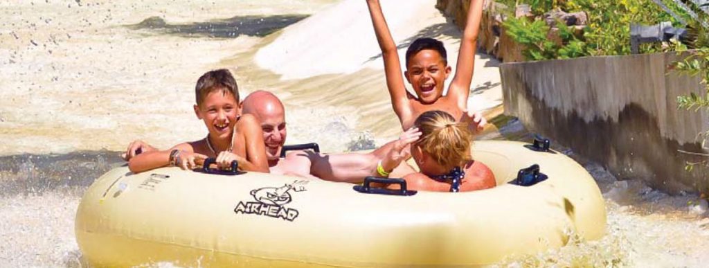 Image of a family going down the water ride in a big yellow tube. Theres a mom and dad along with two young boys who are smiling and facing the camera, one even has his arms in the air.