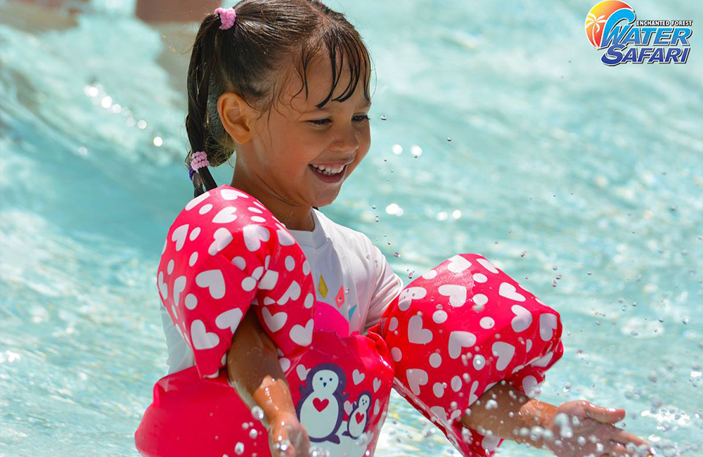 Image of a young girl with pink floats on with hearts and penguins on them. She is looking down smiling at the water as it comes up in a splash.