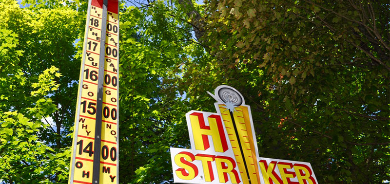 An image of the Hi-Striker arcade game with trees in the background. The yellow and red sign reads "HI STRIKER" and the meter next to it increases in values from 14 to 18.