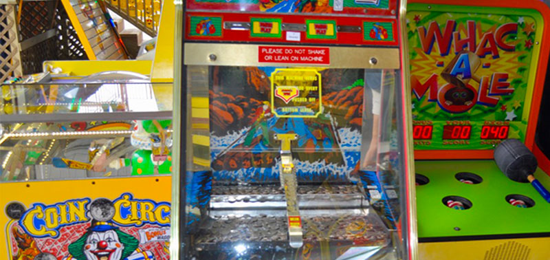 Image of three colorful arcade games at Enchanted Forest Water Safari. The game to the left is called "Coin Circle" and is a yellow machine with an image of a clown on it. The machine in the center has mountain imagery and coins in it. The game on the right is a green machine with a hammer, and is called "Whac A Mole".