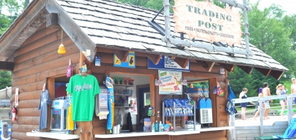 Image of the storefront of the Trading post. You can see a bright green t-shirt hanging up, and then open windows for guests to order from looking into the shop.