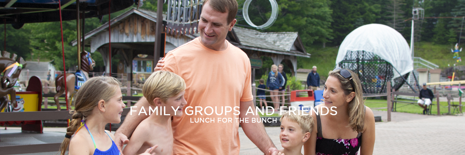 An image of a family walking through the park and smiling. The father, in the middle, is wearing an orange t-shirt. To the right is a young boy and a young girl wearing bathing suits. To the left are a young boy and a woman wearing a bathing suit with sunglasses on her head. Behind them are two wooden structures.