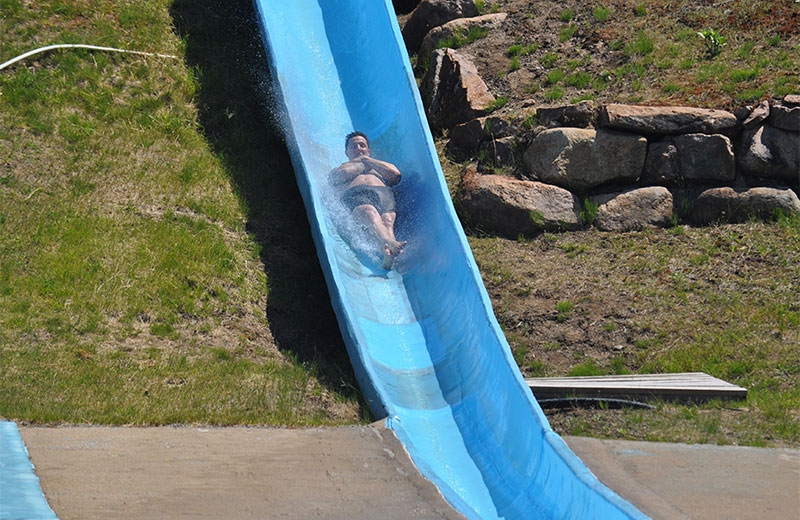 An image of a man lying back with his arms crossed over his chest and feet crossed going down the Killermanajaro water slide. The slide is blue and there are grass and rocks behind it.