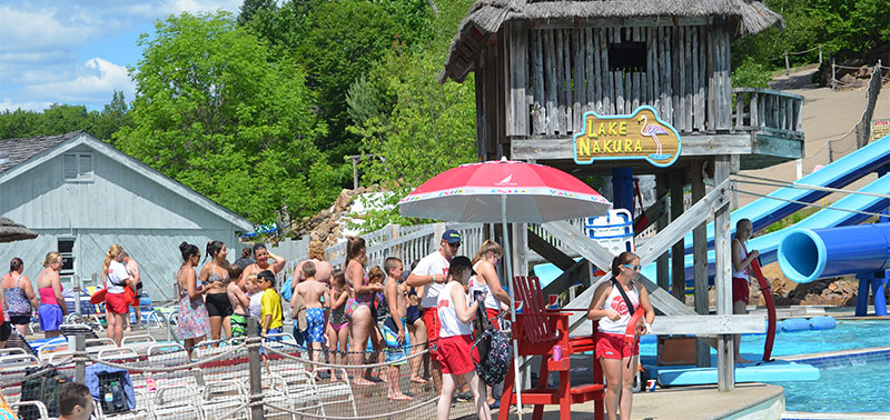 Image of Lake Nakura, a water attraction where you try to cross the pool holding onto a rope. On the left in the image is a crowd of people with beach chairs and to the right is a lifeguard stand with four lifeguards, a wooden structure with a blue and yellow sign reading 'Lake Nakura", and the pool with waterslides visible behind it.