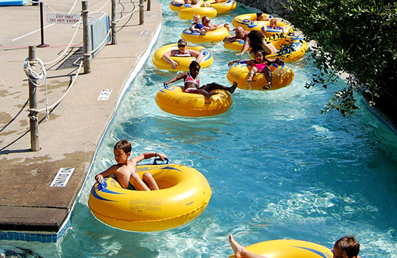 Am image of the Safari River at Enchanted Forest Water Safari. There is a group of people in yellow and blue tubes floating down the river. To the right of the river is greenery, and to the left is pavement with a rope fence around the river.