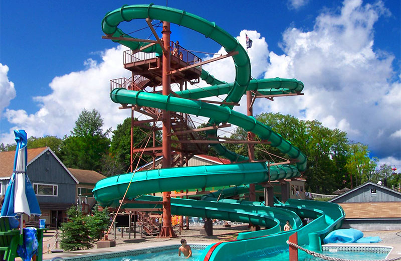 An image of the Shadow waterslides at Enchanted Forest Water Safari. The Shadow slides are green and partially enclosed tubes. The twist around the red stair structure and end in a pool at the bottom. Behind the slides are a blue and brown building. In the front left corner of the image is a blue and white umbrella.