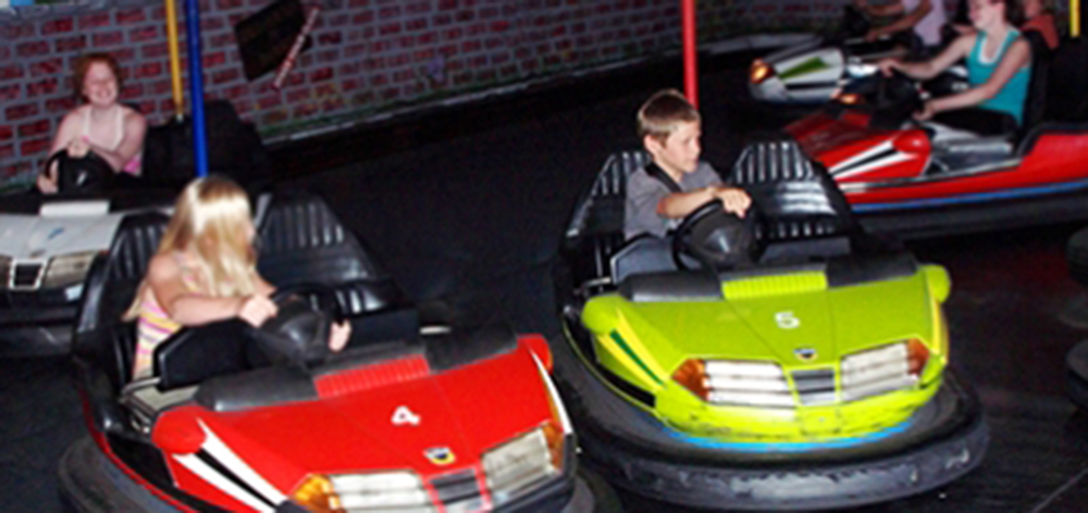 Image of three kids playing on the bumper car rides. One girl is driving a red car trying to hit the boy next to her who is hitting the green car.