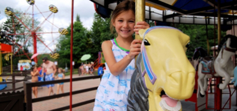 Image of you young girl smiling at the camera as she rides a yellow horse on the carousel.