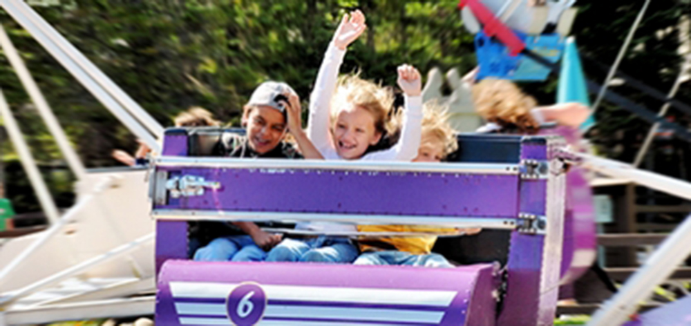 Image of three children on the scrambler ride. There is a little girl in the middle with her hands in the air and a smile on her face.