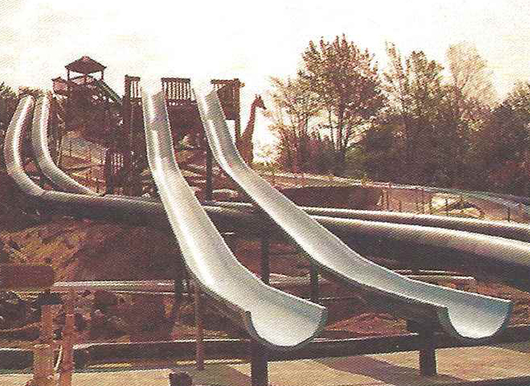 An old image of four water slides at Enchanted Forest Water Safari. Two slides parallel to each other are open air and go straight down. The other two are enclosed and curve beneath the two open-air slides.