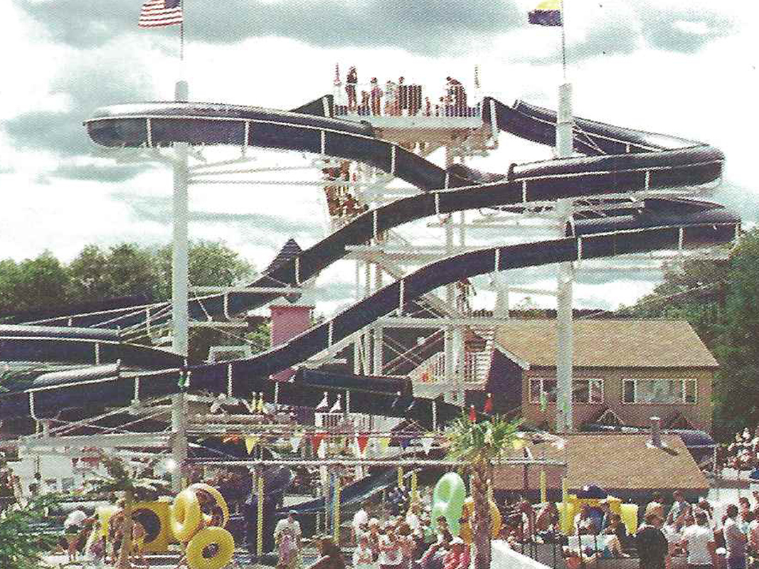 An old image of one of Enchanted Forest Water Safari's original waterslides. The slides are two long, curvy black tubes attached to two white poles with flags at the top of them. There are people lining up a white staircase to ride the slides, Behind the two slides is a brown building. In front of the slides are groups of people standing with green and yellow tubes.