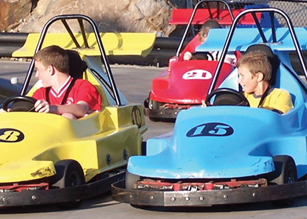 An older image of two young boys riding the Go-Karts at Enchanted Forest Water Safari. The boy on the left is wearing a red t-shirt and driving a yellow car. The boy on the right is wearing a yellow t-shirt and driving a blue car. Behind them is a child driving a red car.