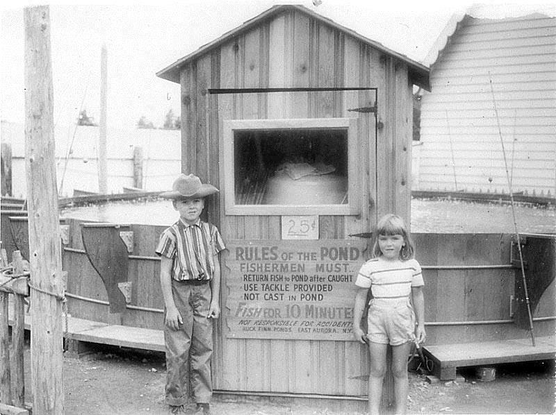 Old photo in black and white of a young boy and girl. They are standing in front of a makeshift catch and release fishing pond.