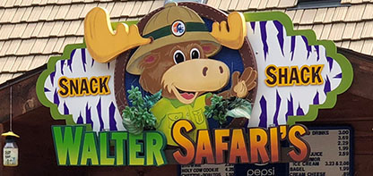 Image of a storefront sign depicting a cartoon moose wearing a hat and green shirt waving with the text Walter Safari's Snack Shack.