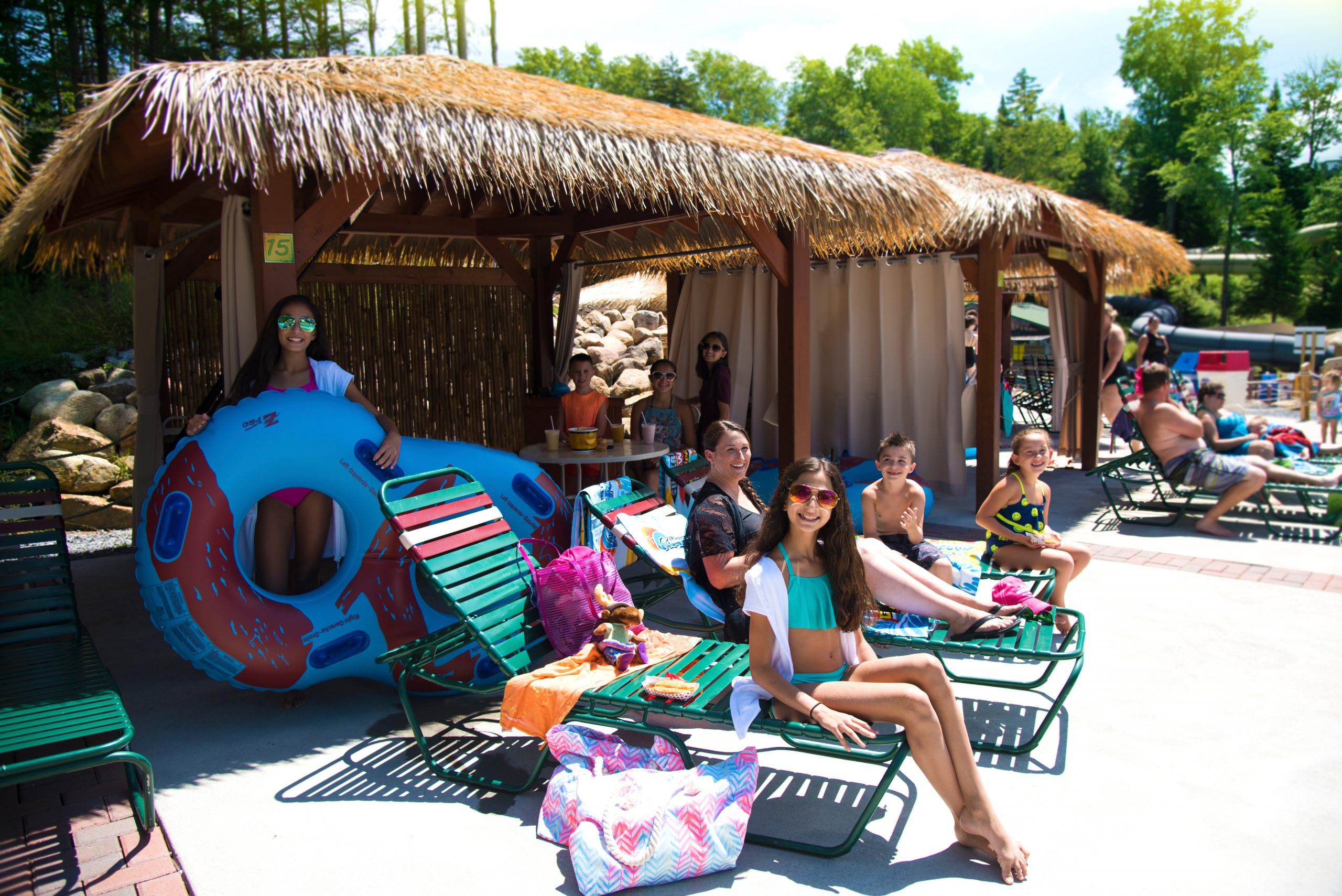 An image of a group of people on beach chairs with towels, tubes and drinks beneath a cabana.