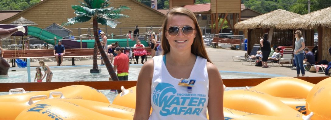 Image of a staff member, infant of yellow tubes, she is smiling at the camera wearing white shirt that says Enchanted Forest Water Safari in blue.