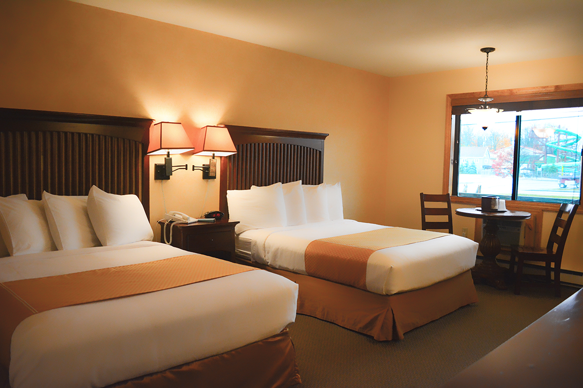 An image of the Standard Double Room at the Water's Edge Inn. There are two beds with white and gold bedding with a table and two lamps between them, and a brown table with two chairs next to the window. Enchanted Forest Water Safari can be seen out the window.