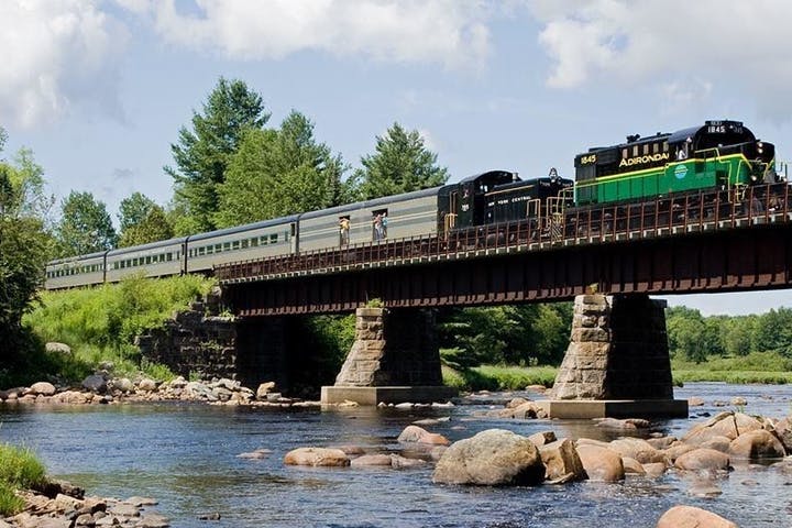 The Adirondack scenic railroad on a bridge over water with boulders sticking out on a sunny day with green trees in the background