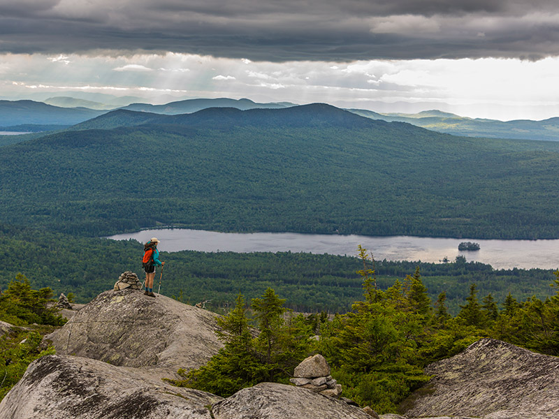 A hiker on top of bald mountain looking out at the lake and trees with the mountains in the distance